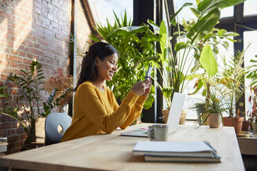 Smiling businesswoman using smart phone sitting at desk in loft office - BSZF02309