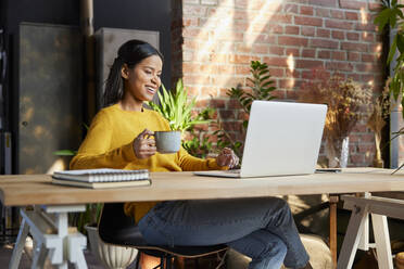 Happy businesswoman with cup doing video call on laptop at loft office - BSZF02312