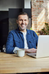 Smiling freelancer with laptop and coffee cup at desk in loft office - BSZF02318