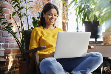 Happy woman using laptop sitting on chair at home - BSZF02322
