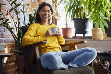 Happy woman holding coffee cup sitting cross-legged in chair at home - BSZF02324