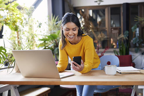 Smiling businesswoman using mobile phone leaning on desk at loft office - BSZF02326