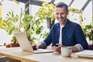 Happy businessman text messaging through smart phone at desk in loft office - BSZF02331