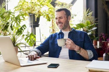 Glücklicher Geschäftsmann mit Laptop und Kaffee im Loft-Büro - BSZF02333