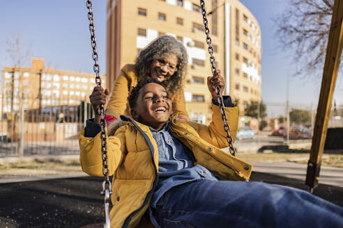 Verspielte ältere Frau, die ihren Enkel auf einer Schaukel auf dem Spielplatz schiebt - JCCMF09919