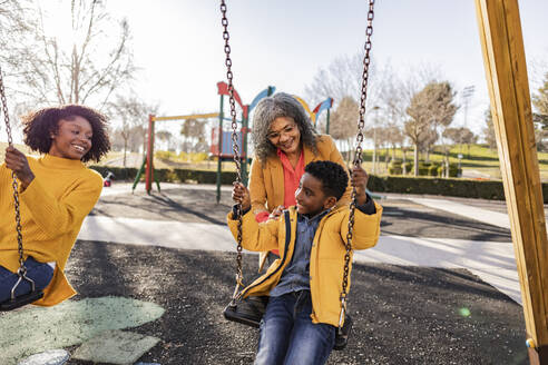 Lächelnde Frau sieht Großmutter an, die ihren Sohn auf dem Spielplatz schaukeln lässt - JCCMF09920