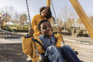 Glückliche Mutter, die ihren Sohn auf dem Spielplatz schaukeln lässt - JCCMF09927