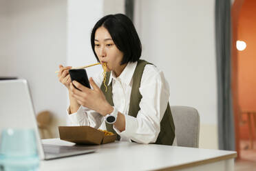 Businesswoman eating noodles and using smart phone in office - EBSF02944