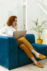 Thoughtful businesswoman sitting with laptop on sofa in office - EBSF02954