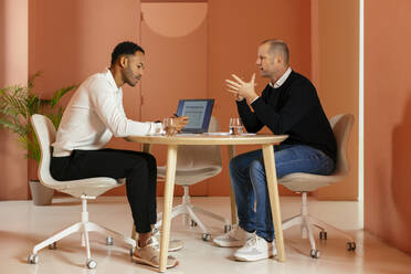 Businessman discussing with colleague at desk in front of orange wall - EBSF02963