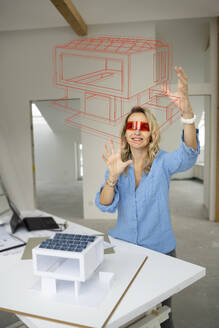 Lächelnder reifer Architekt, der einen Virtual-Reality-Simulator trägt, gestikuliert und ein Hausmodell begutachtet - HMEF01593