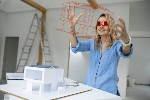 Älterer Architekt, der einen Virtual-Reality-Simulator trägt, gestikuliert und prüft ein Hausmodell auf der Baustelle - HMEF01595