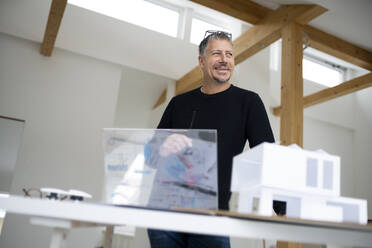 Smiling architect with futuristic tablet PC and model home standing at desk - HMEF01620