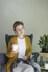 Thoughtful woman holding mug sitting with cat in armchair at home - SVKF01346