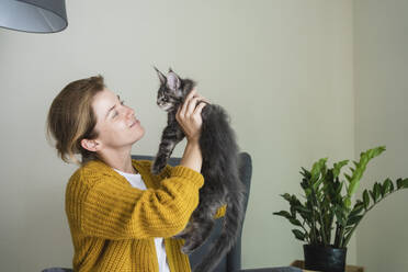 Smiling woman picking up cat near wall at home - SVKF01351