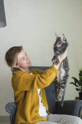 Smiling woman holding cat sitting in armchair at home - SVKF01352