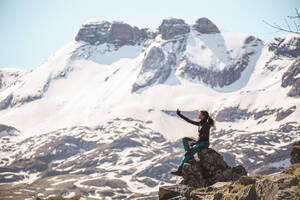 Young woman taking selfie sitting on rock in front of mountains - PCLF00259