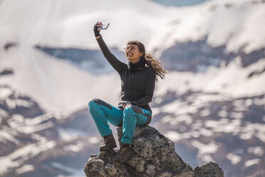 Smiling hiker taking selfie through smart phone sitting on rock - PCLF00260