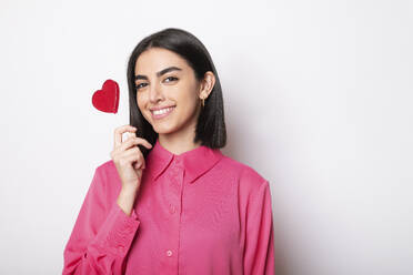 Happy young woman holding red heart shaped lollipop against white background - LMCF00250