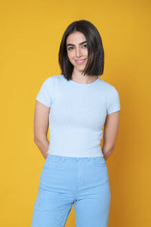 Smiling young woman with hands behind back standing against yellow background - LMCF00258