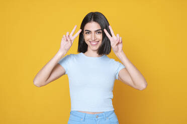 Smiling young woman gesturing peace sign against yellow background - LMCF00261