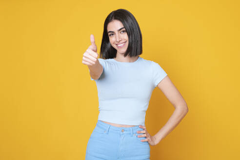 Happy woman showing thumbs up gesture against yellow background - LMCF00262