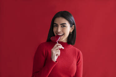 Happy young woman eating heart shaped lollipop against red background - LMCF00285