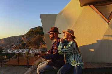 Man playing guitar sitting with girlfriend near eco home at sunset - SYEF00269