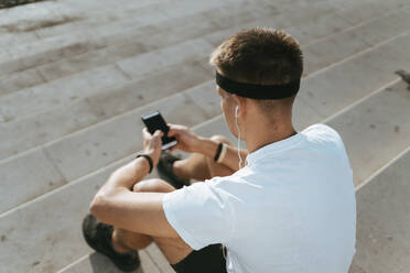 Junger Mann mit Stirnband, der auf einer Treppe Musik über Kopfhörer hört - ANNF00083