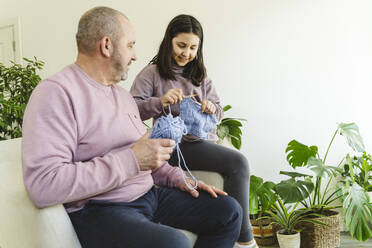 Smiling senior man assisting granddaughter in knitting sweater at home - OSF01443