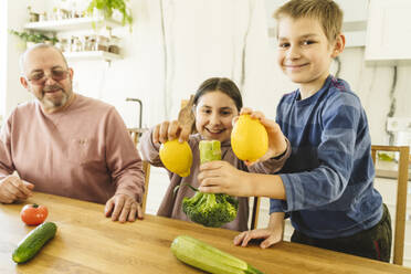 Glücklicher Großvater mit Enkel und Enkelin mit Gemüse am Esstisch - OSF01446