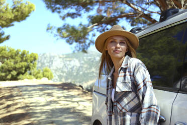 Young woman wearing hat leaning on car - SYEF00273