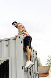 Man climbing on container home in front of clear sky - JJF00503