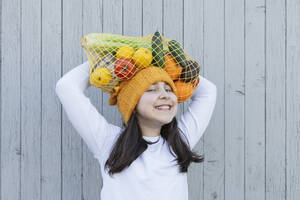 Lächelndes Mädchen, das eine Tüte mit Lebensmitteln auf dem Kopf trägt, vor einer Wand - OSF01466