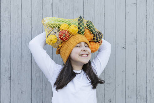 Smiling girl carrying bag of groceries on head in front of wall - OSF01466