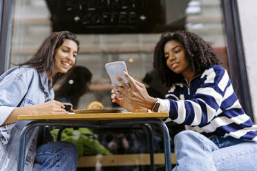 Glückliche junge Frau mit Freund, der sein Smartphone in einem Straßencafé benutzt - JJF00511