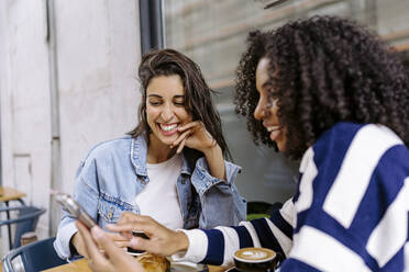 Glückliche junge Frau mit Freund, der in einem Straßencafé sein Handy benutzt - JJF00514