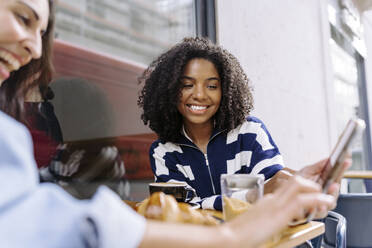 Glückliche junge Frau, die mit einem Freund in einem Café ein Mobiltelefon benutzt - JJF00525