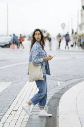Junge Frau mit Tragetasche und Smartphone beim Überqueren der Straße - JJF00535