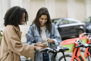 Glückliche Frau mit Freund beim Bezahlen per Smartphone an einer Fahrradparkstation - JJF00538