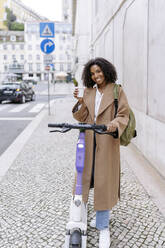 Fröhliche junge Frau mit Kaffeetasse und Elektroroller auf dem Fußweg stehend - JJF00549