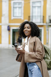 Happy young woman with smart phone and coffee cup standing in front of building - JJF00577