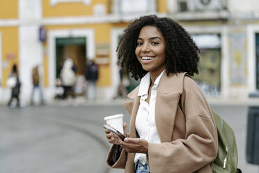 Happy young woman standing with coffee cup and mobile phone - JJF00579