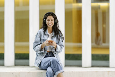 Happy woman wearing wireless headphones sitting with smart phone in front of building - JJF00618