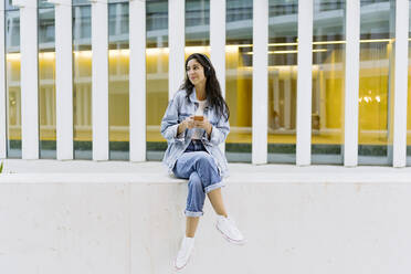 Lächelnde junge Frau mit Smartphone an der Wand sitzend - JJF00619