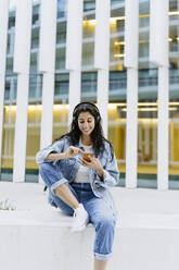Lächelnde junge Frau mit Smartphone an der Wand sitzend - JJF00621