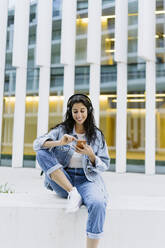 Smiling young woman using smart phone sitting on wall - JJF00621