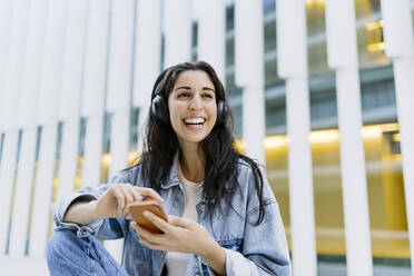 Glückliche junge Frau mit Smartphone vor einem Gebäude - JJF00622