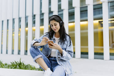 Lächelnde Frau mit Smartphone an der Wand vor einem Gebäude sitzend - JJF00623