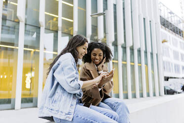 Happy young woman with friend using smart phone in front of building - JJF00627